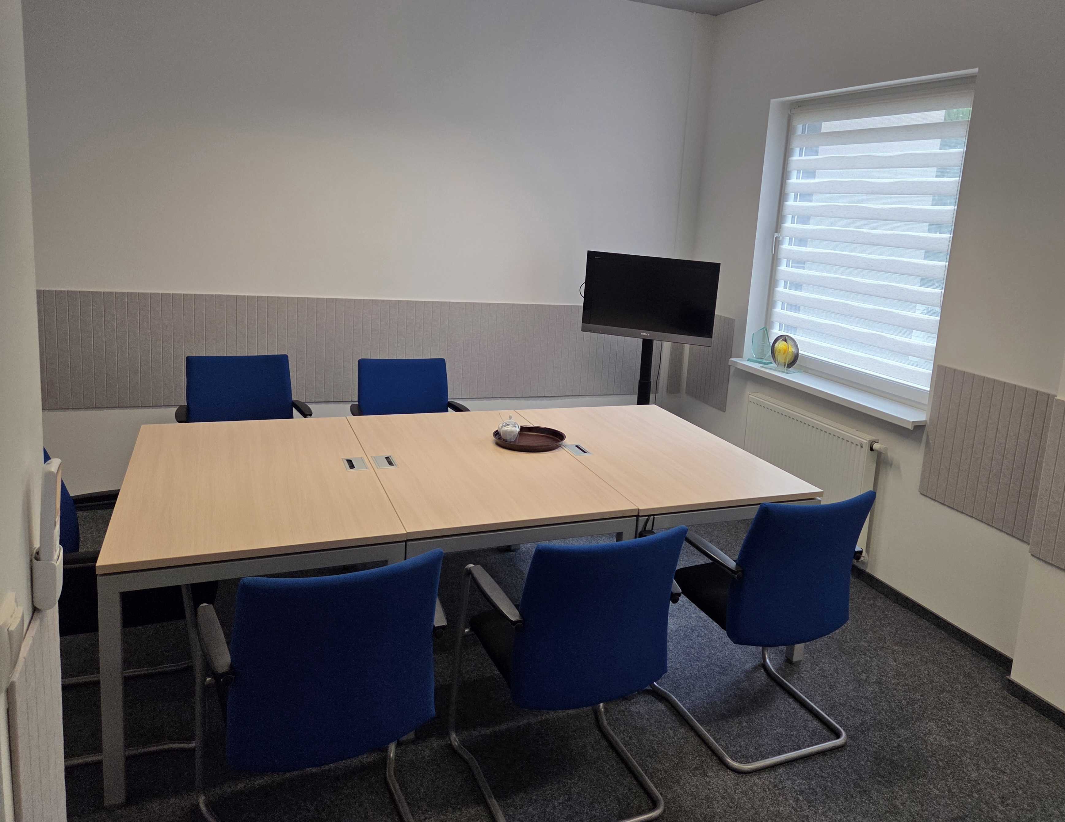 Zdjęcie sali konferencyjnej z nowymi stołami, krzesłami, telewizorem na stojaku i nagrodami na parapecie.