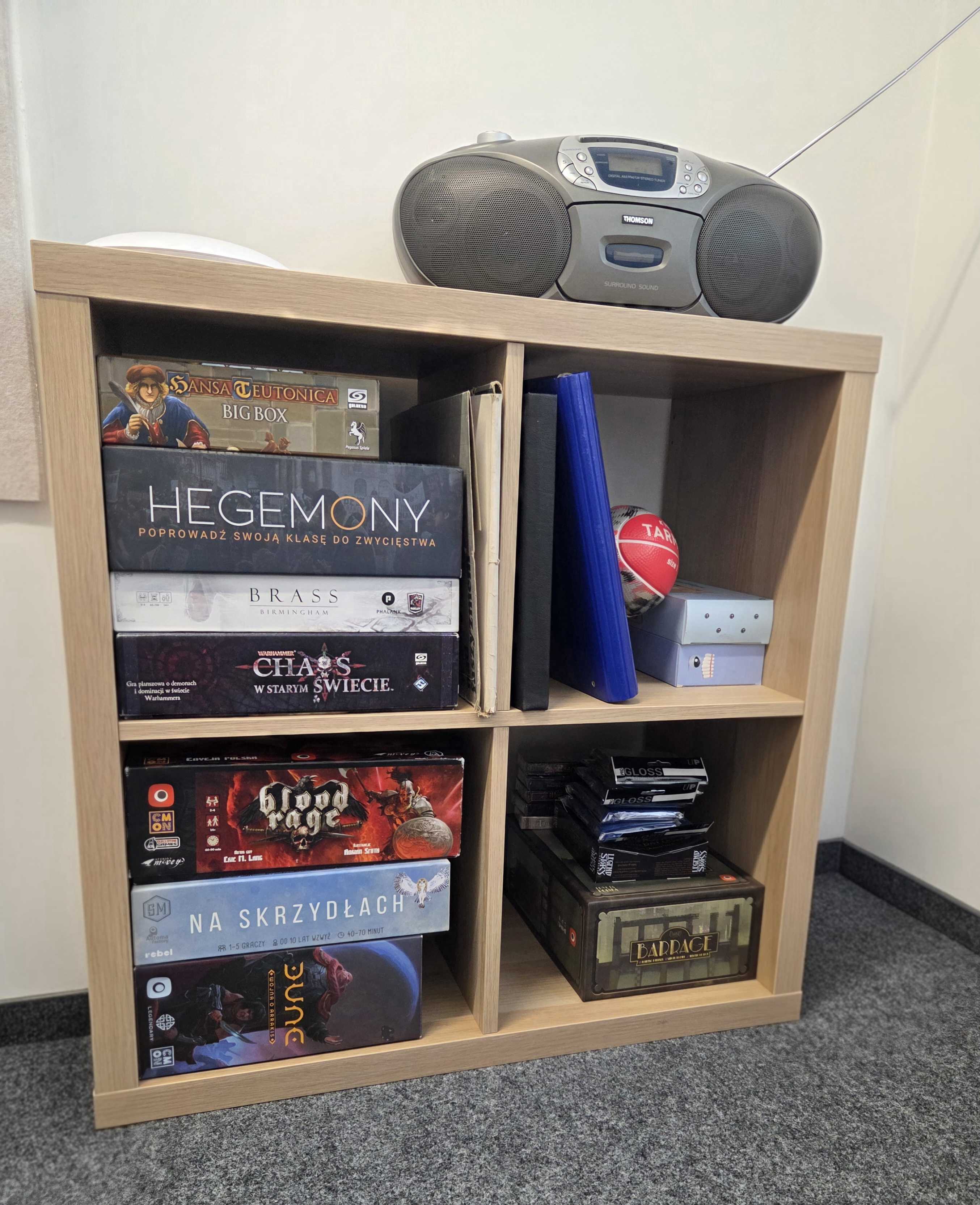 Zdjęcie szafki, na której widać radio i gry planszowe oraz karciane, m.in. Hegemony, Hansa Teutonica, Brass Birmingham, Warhammer Chaos w Starym Świecie, Blood Rage, Na Skrzydłach, Dune, Barrage i Flesh and Blood.