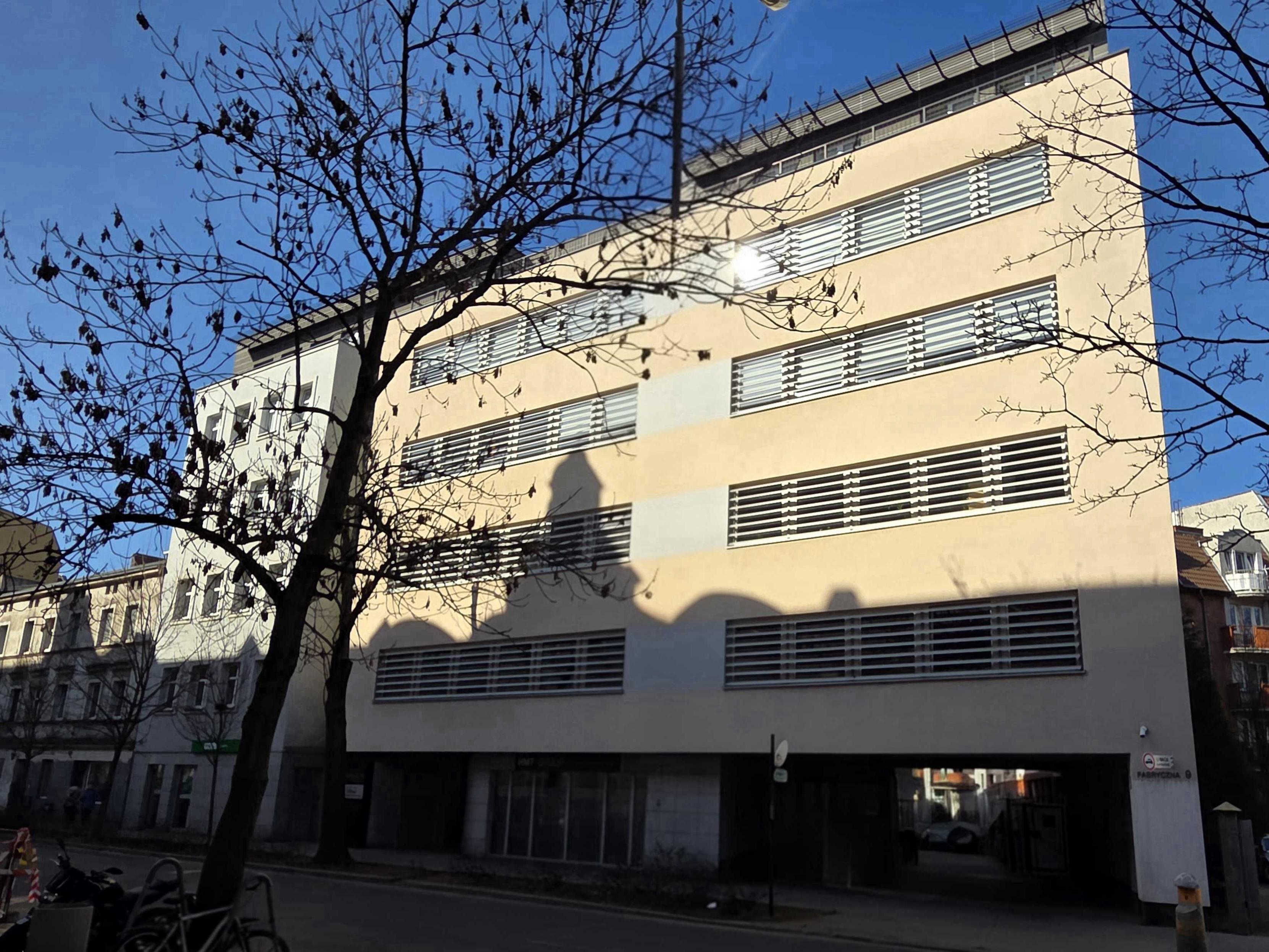 Zdjęcie nowej siedziby Wildy Software - jasnego biurowca na Fabrycznej 9, na tle drzewa, którego cień bada na budynek.