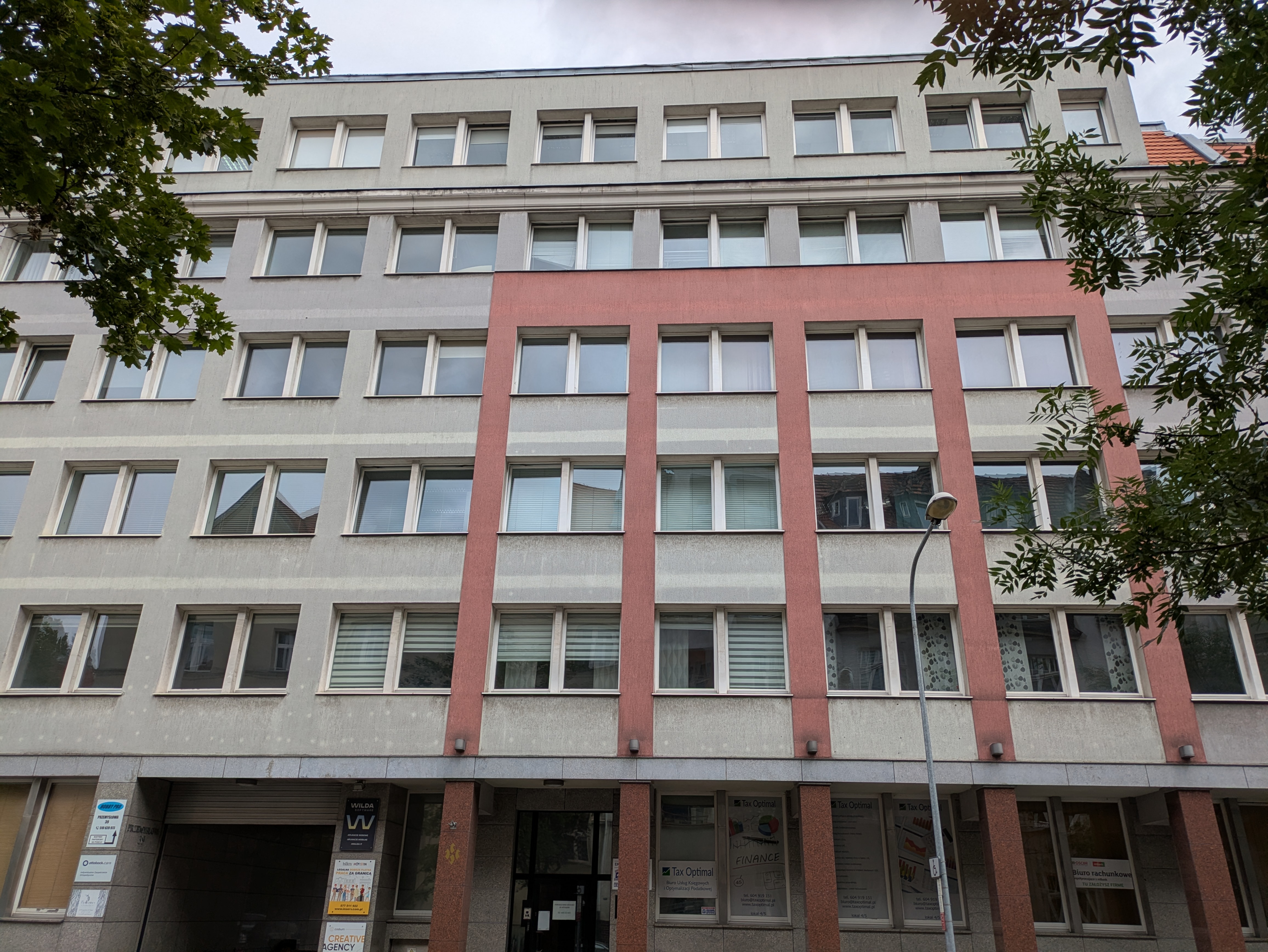 Zdjęcie z dołu pokazujące budynek biurowy składający się z pięciu pięter, gdzie na górze są otwarte okna - to stare biuro Wilda Software. Budynek jest szary z czerwonymi wstawkami.