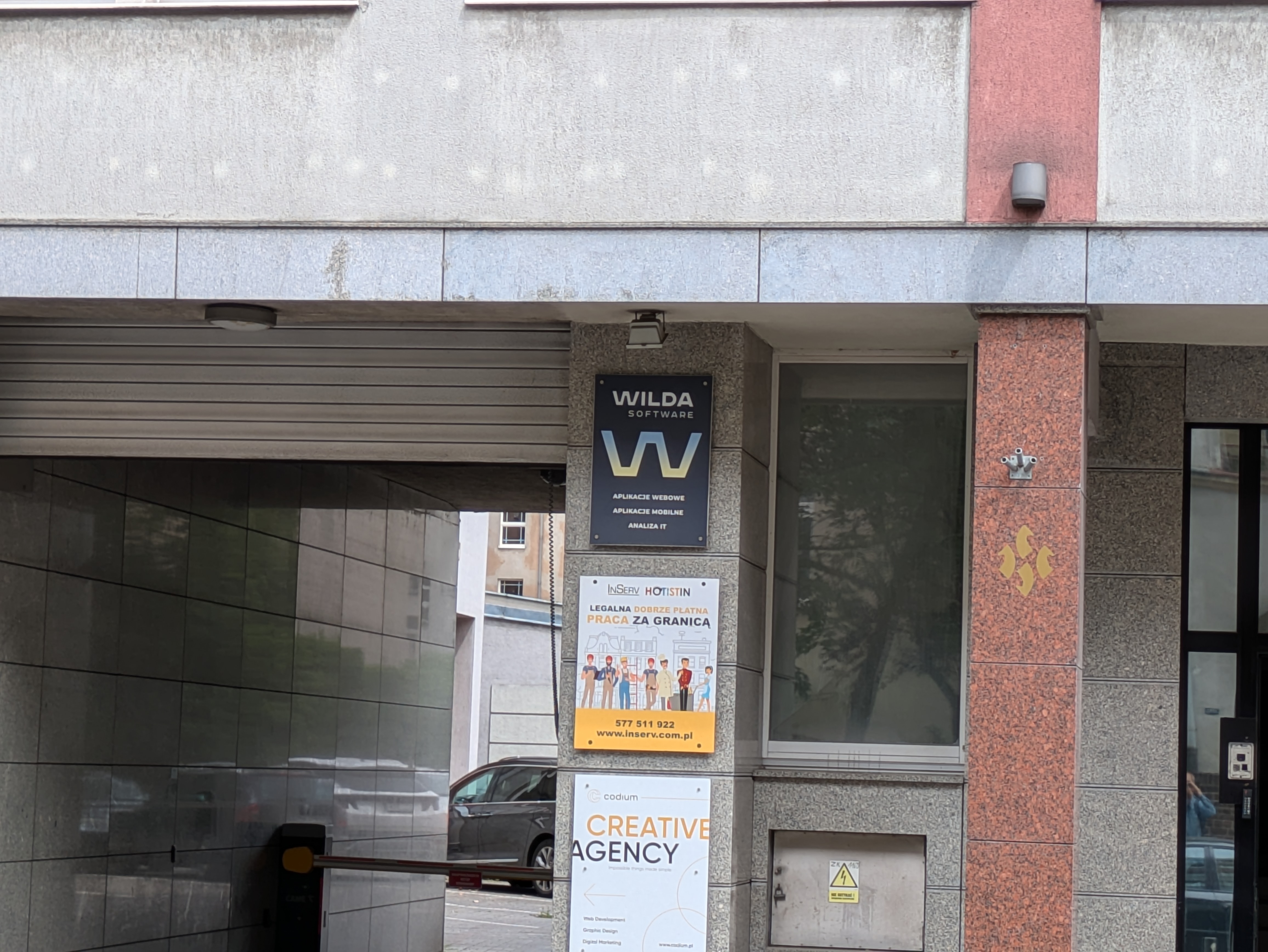 Szyld Wilda Software widoczny na ścianie budynku, gdzie w oddali widać szlaban i parking. Pod spodem są szyldy innych firm, a ściana należy do budynku na Przemysłowej 39.